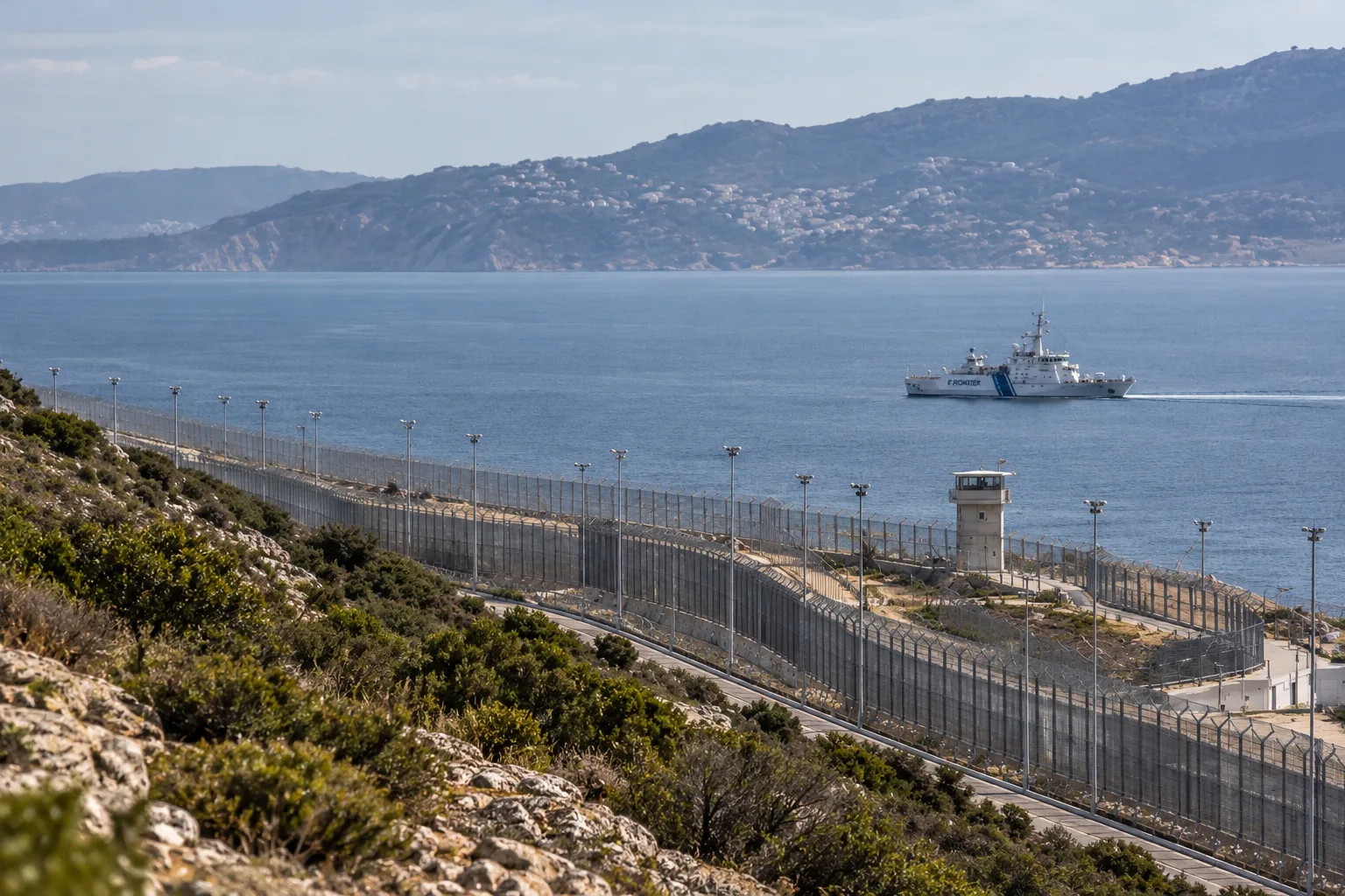 Blick auf einen gesicherten Abschnitt der EU-Außengrenze mit Grenzzaun, Wachturm und einem Frontex-Patrouillenschiff auf dem Meer. Symbolbild zum Ausbau von Frontex.