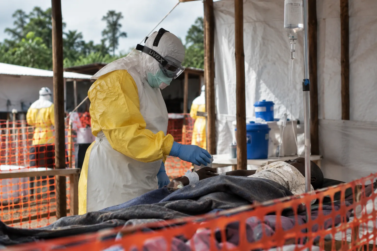 Eine medizinische Fachkraft in vollständiger Schutzkleidung behandelt einen Patienten in einer Isolierstation. Im Hintergrund sind weitere Behandlungszelte und Einsatzkräfte zu sehen. Symbolbild zum Ebola-Ausbruch im Kongo.