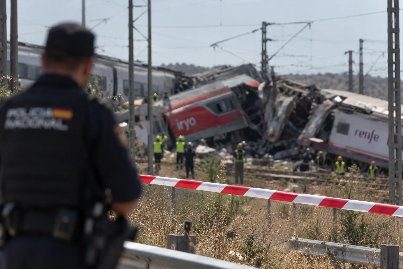 Rot-weißes Absperrband im Vordergrund einer Zugunfallstelle. Dahinter stehen entgleiste Hochgeschwindigkeitszüge von Iryo und Renfe sowie Einsatzkräfte auf den Gleisen. Ein unscharfer Beamter der spanischen Policía Nacional blickt auf die Unfallstelle.