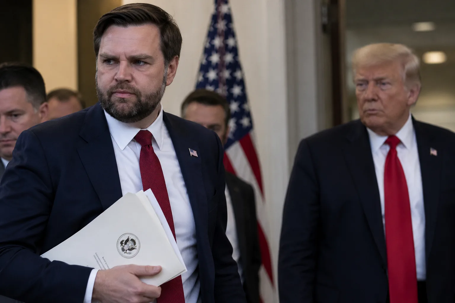 US-Vizepräsident JD Vance mit einer Dokumentenmappe im Vordergrund, während Donald Trump unscharf im Hintergrund zu sehen ist. Das Bild symbolisiert die widersprüchlichen Angaben zu einem möglichen Schreiben an den Iran.